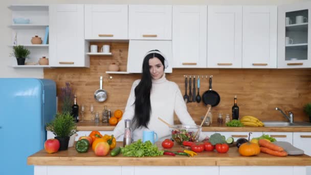 Retrato de una joven morena usando auriculares cocinando en la cocina . — Vídeo de stock