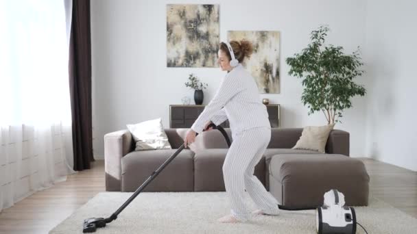 Mujer feliz limpiando el hogar, bailando con aspiradora usando pijamas y auriculares . — Vídeos de Stock