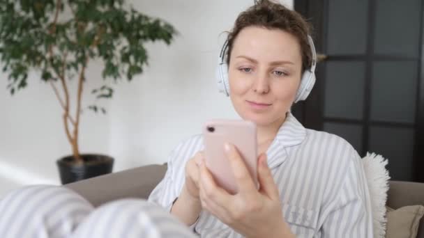 Retrato de mujer joven usando Smartphone y usando auriculares relajantes en casa — Vídeos de Stock