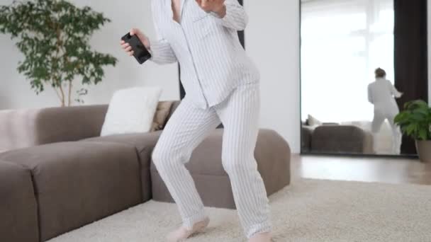 Veselá dívka ve sluchátkách tančí s mobilním telefonem v obývacím pokoji. — Stock video