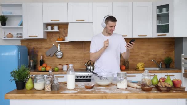 Hombre joven feliz en camiseta blanca bailando con Smartphone en auriculares inalámbricos en la cocina . — Vídeo de stock