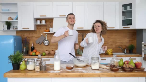 Casal jovem com copos de café dançando juntos na cozinha enquanto cozinha café da manhã . — Vídeo de Stock