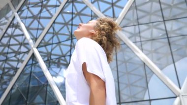 Young Stylish Woman In Wireless Earphones Listening Music Against Modern City Architecture