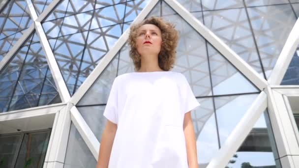 Νεαρή κομψή γυναίκα που περπατά σε λευκό φόρεμα στην πόλη με τη σύγχρονη αρχιτεκτονική — Αρχείο Βίντεο