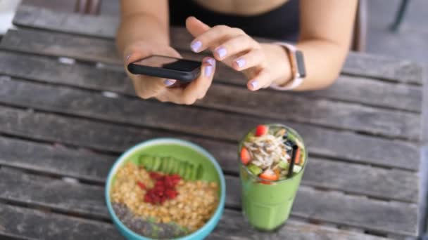 Kadın cep telefonu kullanıyor ve kahvaltıda sağlıklı meyve suyu kasesi yiyor. — Stok video
