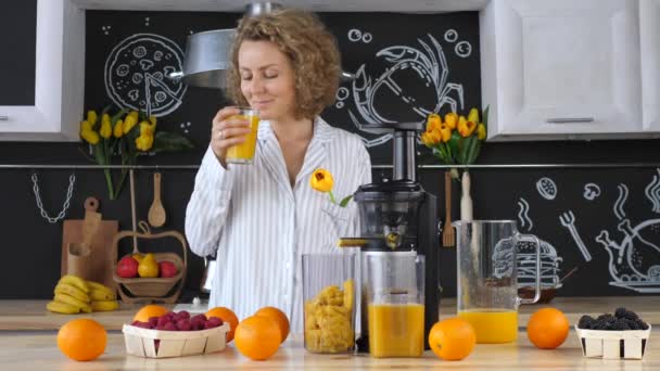 Jeune femme blonde en pyjama boire du jus d'orange frais dans la cuisine — Video