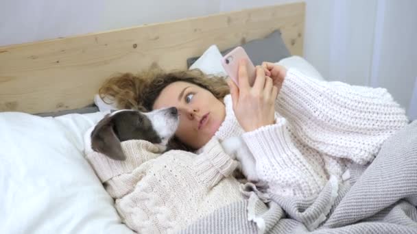 Coziness, estilo de vida, tecnología, gente y concepto de la mascota. Chica usando teléfono celular con perro acostado en la cama . — Vídeos de Stock