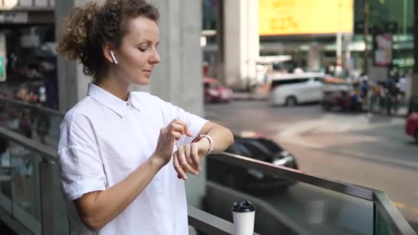 Mujer usando smartwatch en auriculares inalámbricos en la ciudad — Vídeos de Stock