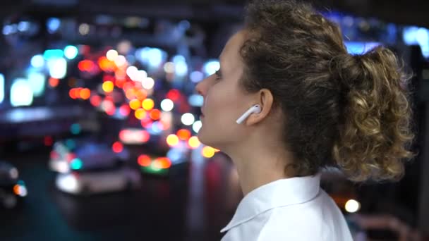 Menina vestindo fones de ouvido sem fio Bluetooth contra Bokeh Cidade Noite — Vídeo de Stock