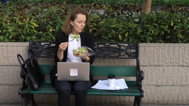 Egy üzletasszony laptoppal ebédel a parkban, telefonon beszél Airpodsban. — Stock videók