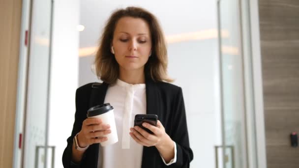 Mujer joven de negocios con café usando teléfono inteligente caminando en el pasillo de la oficina — Vídeos de Stock