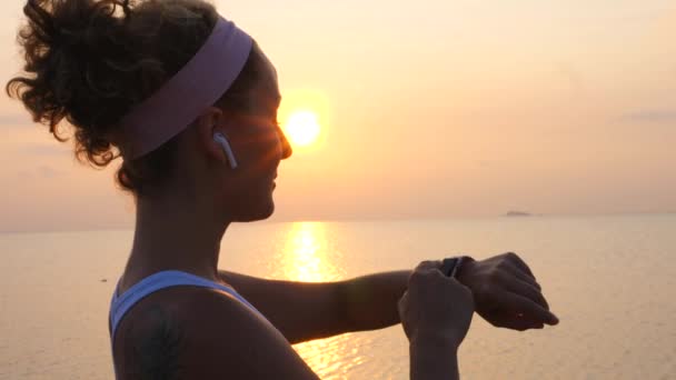 Sport actif femme dans les écouteurs sans fil en utilisant Smartwatch au coucher du soleil sur la plage — Video