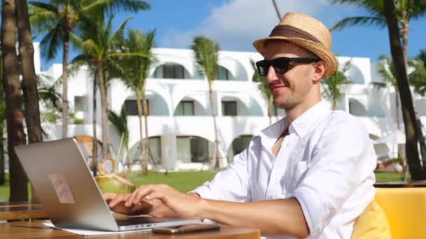 Digital Nomad Man In Hat работает удаленно на ноутбуке на пляже . — стоковое видео