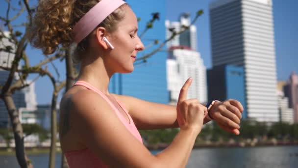 Jonge vrouw in draadloze oortelefoons met behulp van Smartwatch na het joggen buiten — Stockvideo