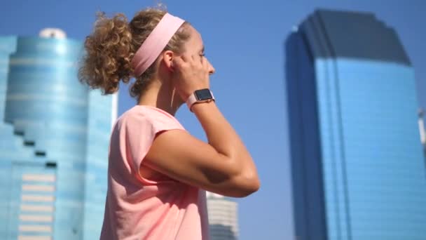 Молодая спортивная женщина слушает музыку в беспроводных наушниках с умными часами в городе — стоковое видео
