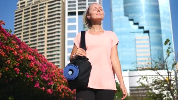 Nő sétál a City Park jóga matrac hallgat zenét vezeték nélküli fülhallgató — Stock videók