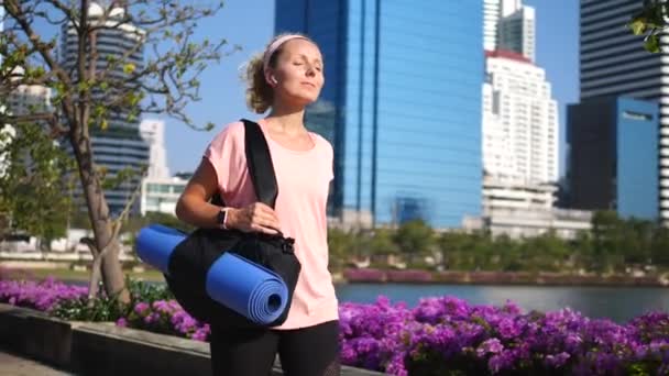 Fitness kobieta spaceru w parku miejskim z Yoga Mat noszenie słuchawki bezprzewodowe — Wideo stockowe