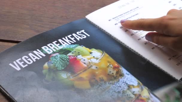 Primer plano de la mano femenina la elección de desayuno vegano en el menú del restaurante — Vídeo de stock