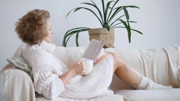 Mujer sentada en un sofá, bebiendo té y leyendo un libro. Ambiente doméstico acogedor . — Vídeos de Stock