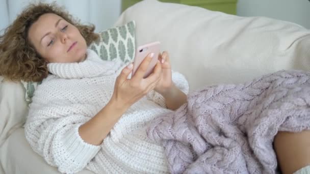 Chica con el teléfono celular acostado en el sofá debajo de acogedora manta caliente — Vídeos de Stock