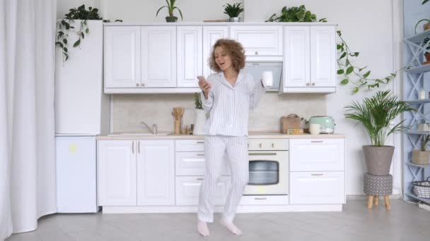 Młoda podekscytowana kobieta taniec w piżamie gospodarstwa smartfon w kuchni. — Wideo stockowe