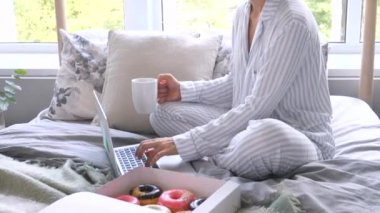 Üzerinde pijama olan bir kadın rahat yatağında dizüstü bilgisayar ve bir fincan kahve kullanıyor.