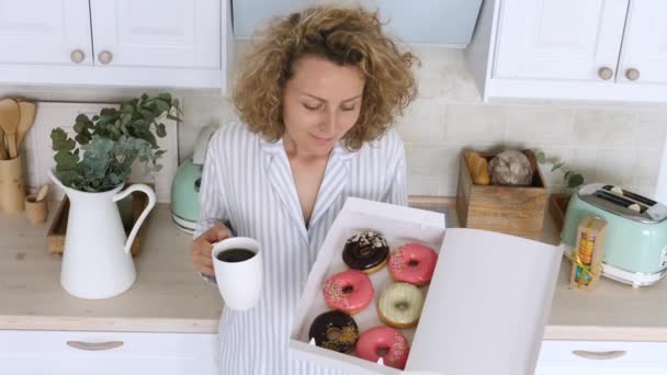 Jonge vrouw draagt pyjama ontbijten met koffie en donut op keuken — Stockvideo