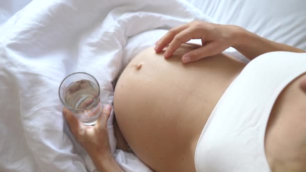 Primo piano della donna incinta che tiene un bicchiere d'acqua e si tocca la pancia a letto . — Video Stock