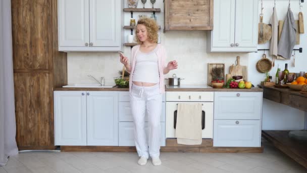 Gelukkig zwanger vrouw met behulp van smartphone en dansen in de keuken. — Stockvideo