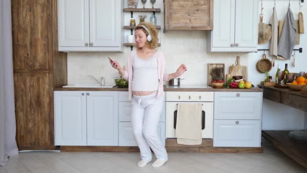 Vacker gravid flicka dansar med smartphone i köket. — Stockvideo