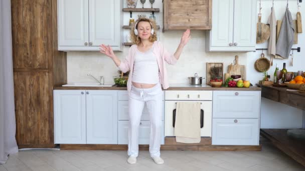 Счастливая беременная женщина в наушниках танцует на кухне . — стоковое видео