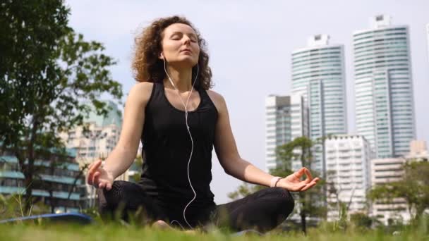 Mujer joven con auriculares haciendo meditación en el parque de la ciudad . — Vídeo de stock