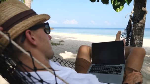 Hombre de negocios joven con el ordenador portátil que se balancea en la hamaca en la playa — Vídeo de stock