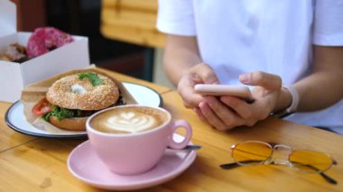 Kadın Elleri Kahve ve simit ile Kahvaltı yaparken Cep Telefonu Kullanıyor