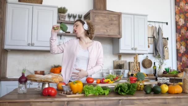 Mooi zwanger vrouw zingen in broccoli en dansen in hoofdtelefoon op keuken. — Stockvideo