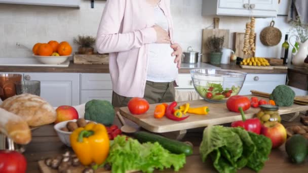 Беременная женщина касается живота во время приготовления здорового салата на кухне . — стоковое видео