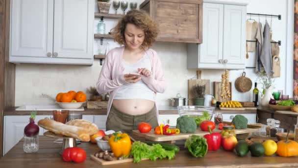 Красивая беременная женщина с помощью смартфона и приготовления пищи на кухне . — стоковое видео