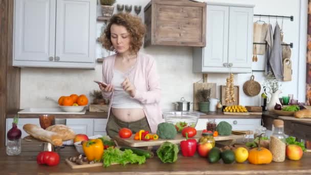 Молодая беременная женщина ищет рецепт на смартфоне и делает салат на кухне . — стоковое видео