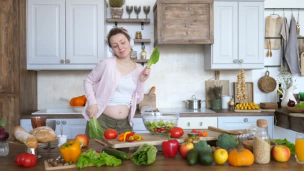 Happy femme enceinte dansant avec des feuilles de salade verte dans la cuisine tout en cuisinant . — Video