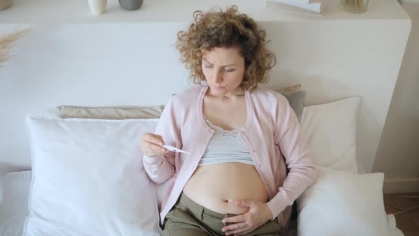 Sorglig sjuk gravid kvinna tar temperatur med termometer på sängen. — Stockvideo