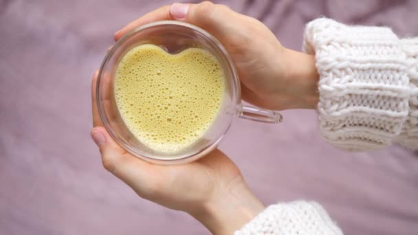 Vrouw handen houden gouden kurkuma amandel latte in glazen beker met hart vorm. — Stockvideo