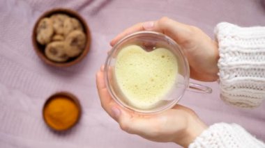 Altın sarısı latte kalp şeklinde cam kupa tutan kadın elleri.