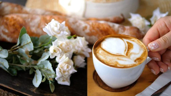 Mano de mujer con taza de café con leche cerca de baguettes y flores — Foto de Stock