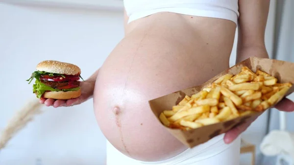 Concepto de antojos de embarazo. Mujer embarazada sosteniendo hamburguesa y patatas fritas . — Foto de Stock