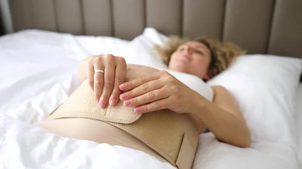 Hamile kadın yatağa uzanıyor ve abdominal destek bandajı takıyor. Annelik Destek Ürünleri. — Stok fotoğraf