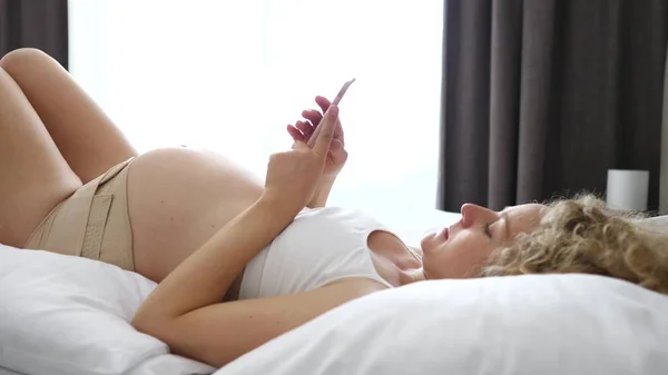 Беременность, материнство, технологии, люди и ожидания Концепция - Беременная женщина со смартфоном в постели . — стоковое фото