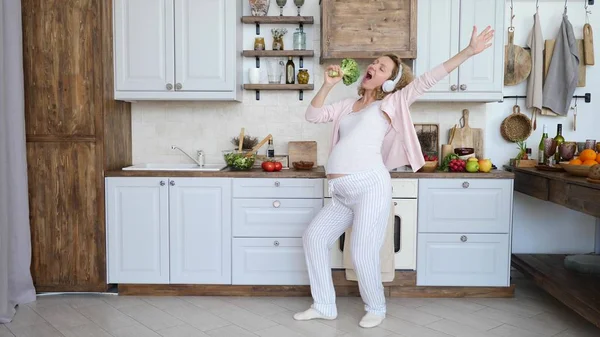 Молодая беременная женщина поет в брокколи в наушниках, танцует на кухне . — стоковое фото