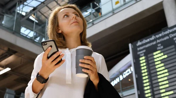 Молода ділова жінка з кавою використовуючи смартфон на рейсі аеропорту Розклад — стокове фото