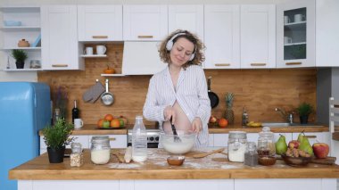 Sağlıklı Hamilelik Konseptin kutlu olsun. Kulaklıklı hamile kadın mutfakta dans ediyor ve yemek pişiriyor..