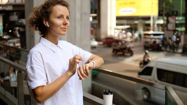 Mujer usando smartwatch en auriculares inalámbricos en la ciudad —  Fotos de Stock